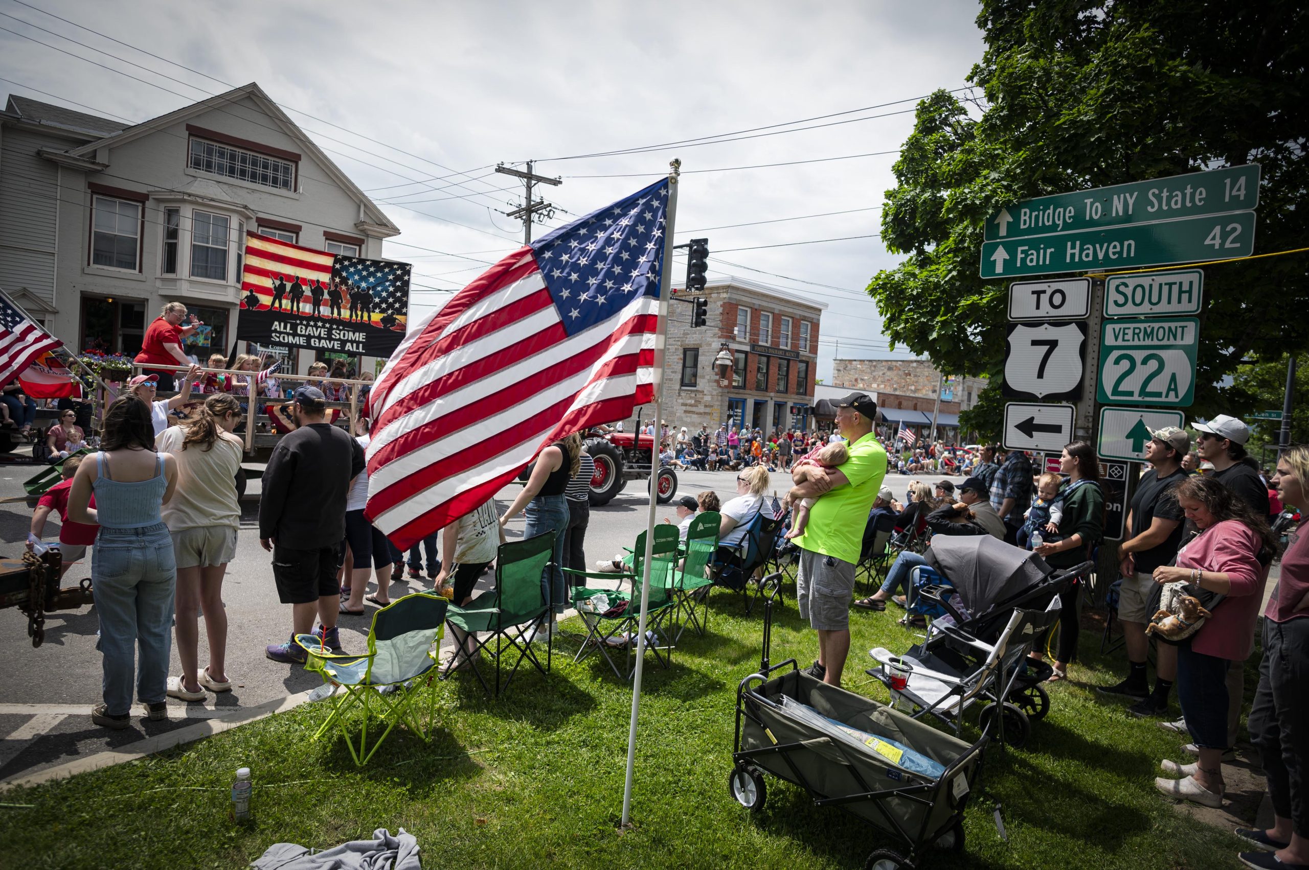 A crowd watches a parade on a street lined with American flags and signposts indicating directions to state routes. A large American flag waves prominently in the foreground.