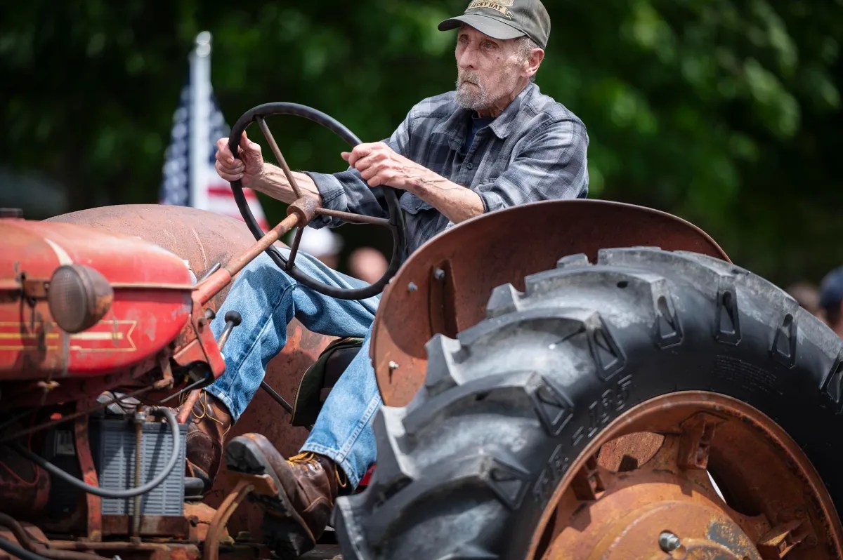 An elderly man drives an old, rusted red tractor while wearing a plaid shirt and cap. An American flag is visible in the background.