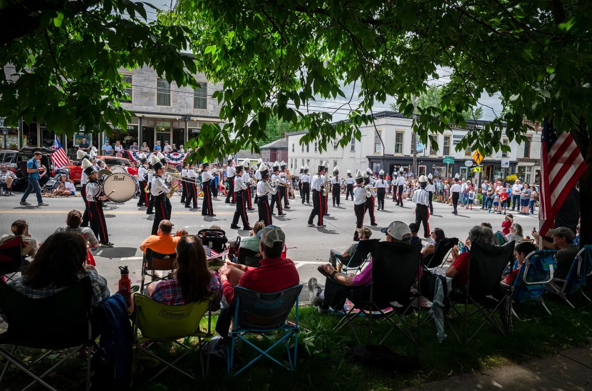 A marching band performs in a street parade as onlookers seated on lawn chairs watch from under shaded trees on a sunny day. American flags are visible among the crowd.
