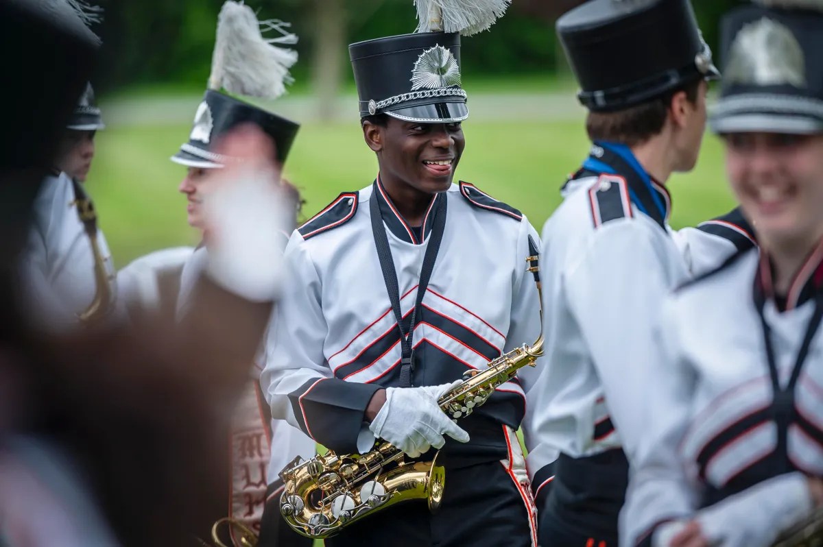 A marching band member in a black and white uniform holds a saxophone and smiles. Others in similar attire are visible in the background. The scene is outdoors with grass and trees.