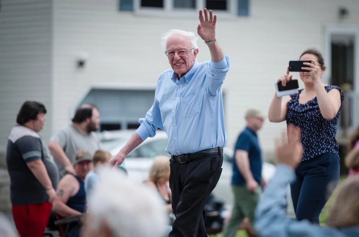 An elderly man with white hair and glasses waves at a group of people outdoors. A woman stands nearby taking a photo with her phone. Other individuals are in the background.