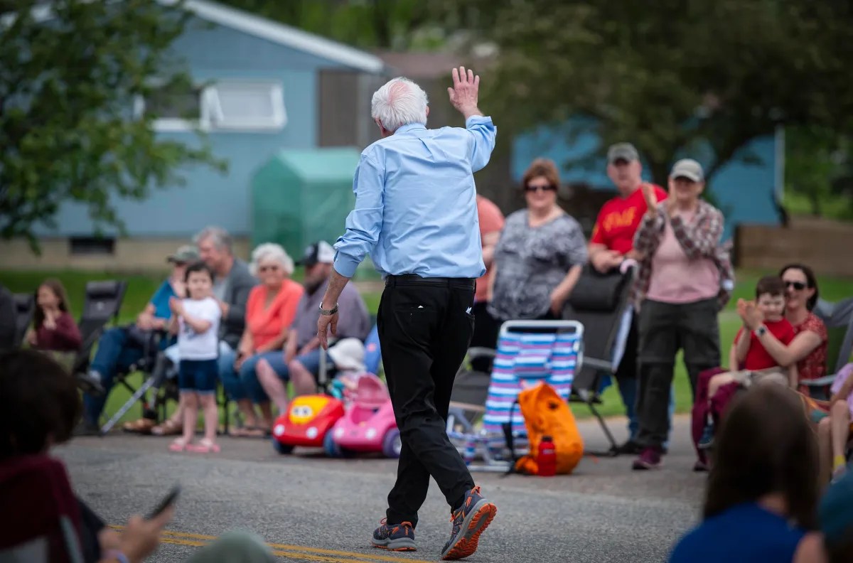 A man in a light blue shirt waves while walking past a seated outdoor audience. Some people clap; there are toys and chairs on the sidewalk.