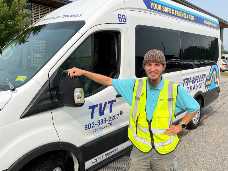 A person wearing a yellow safety vest and beanie stands beside a white "Tri-Valley Transit" van, resting one hand on the driver's side mirror. The van has contact information on the side.