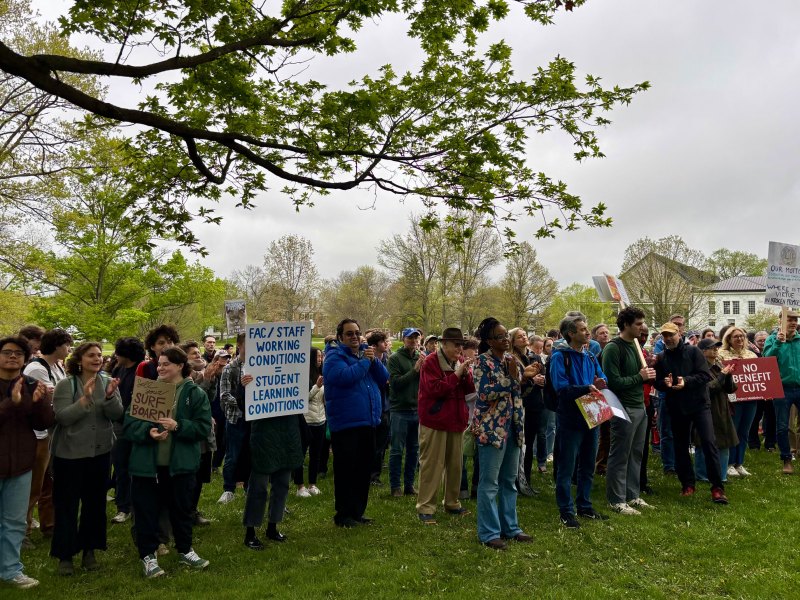 A group of people gather outdoors; several hold signs advocating for faculty, staff, and student conditions and against benefit cuts. Trees and buildings are visible in the background.