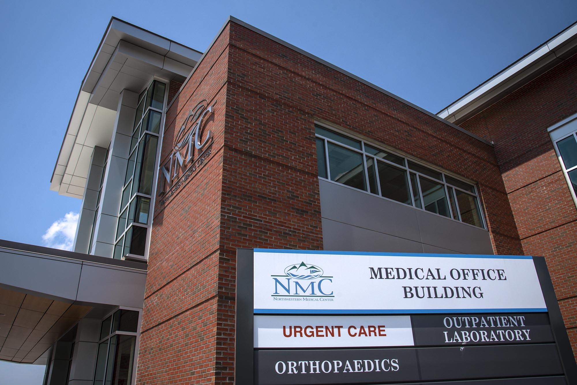Brick medical office building with "NMC" logo and sign listing Urgent Care, Outpatient Laboratory, Orthopaedics. Blue sky in the background.