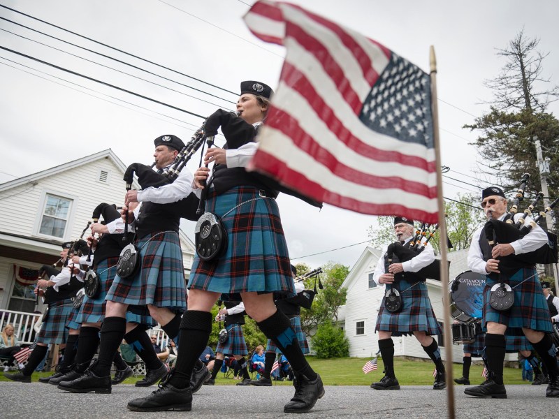 People in matching plaid uniforms play bagpipes while marching in a parade; an American flag is visible in the foreground.