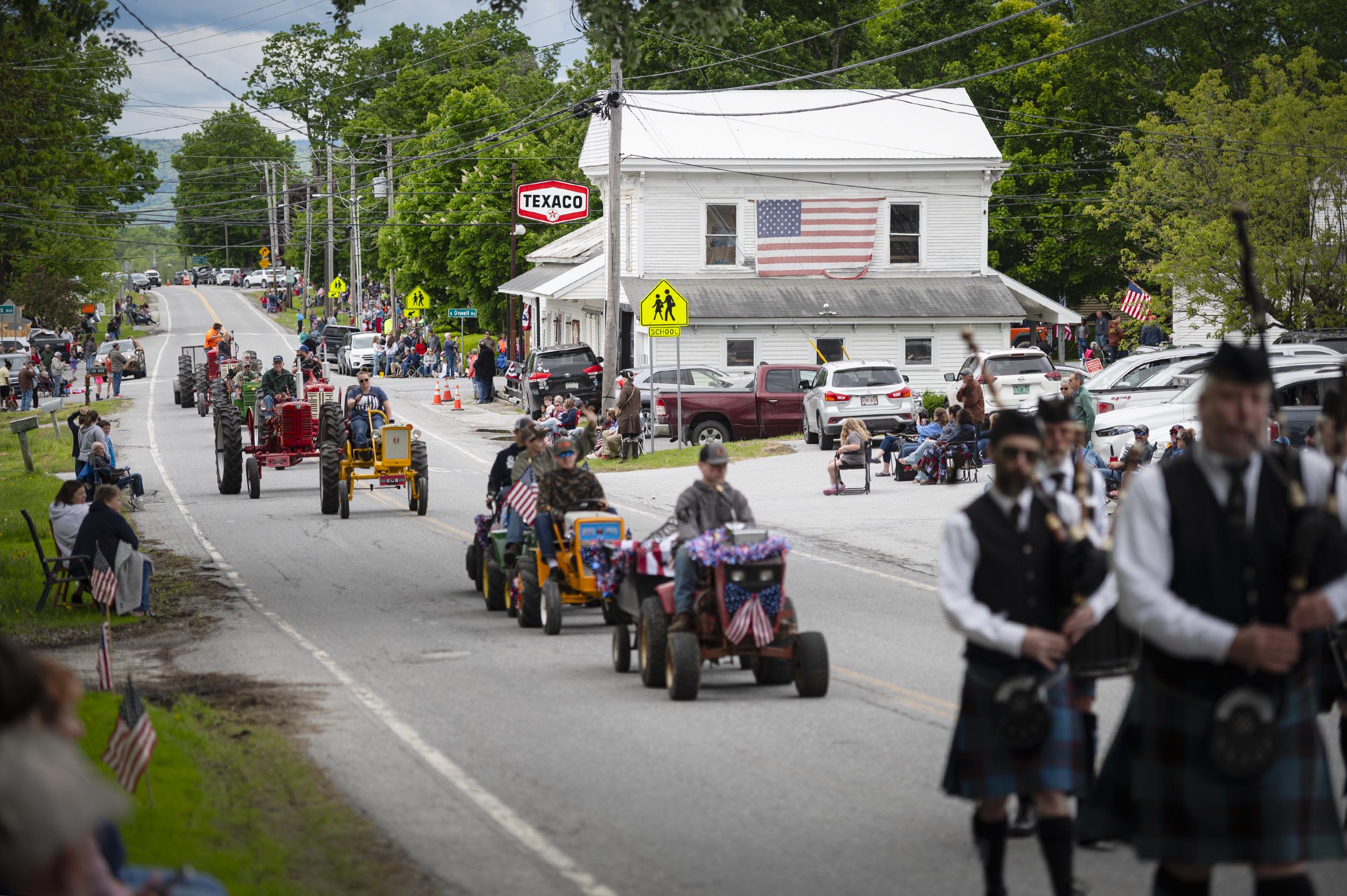 A small-town parade with people watching from sidewalks, tractors driving down the street, and a group of people in kilts marching at the front. An American flag hangs on a building.