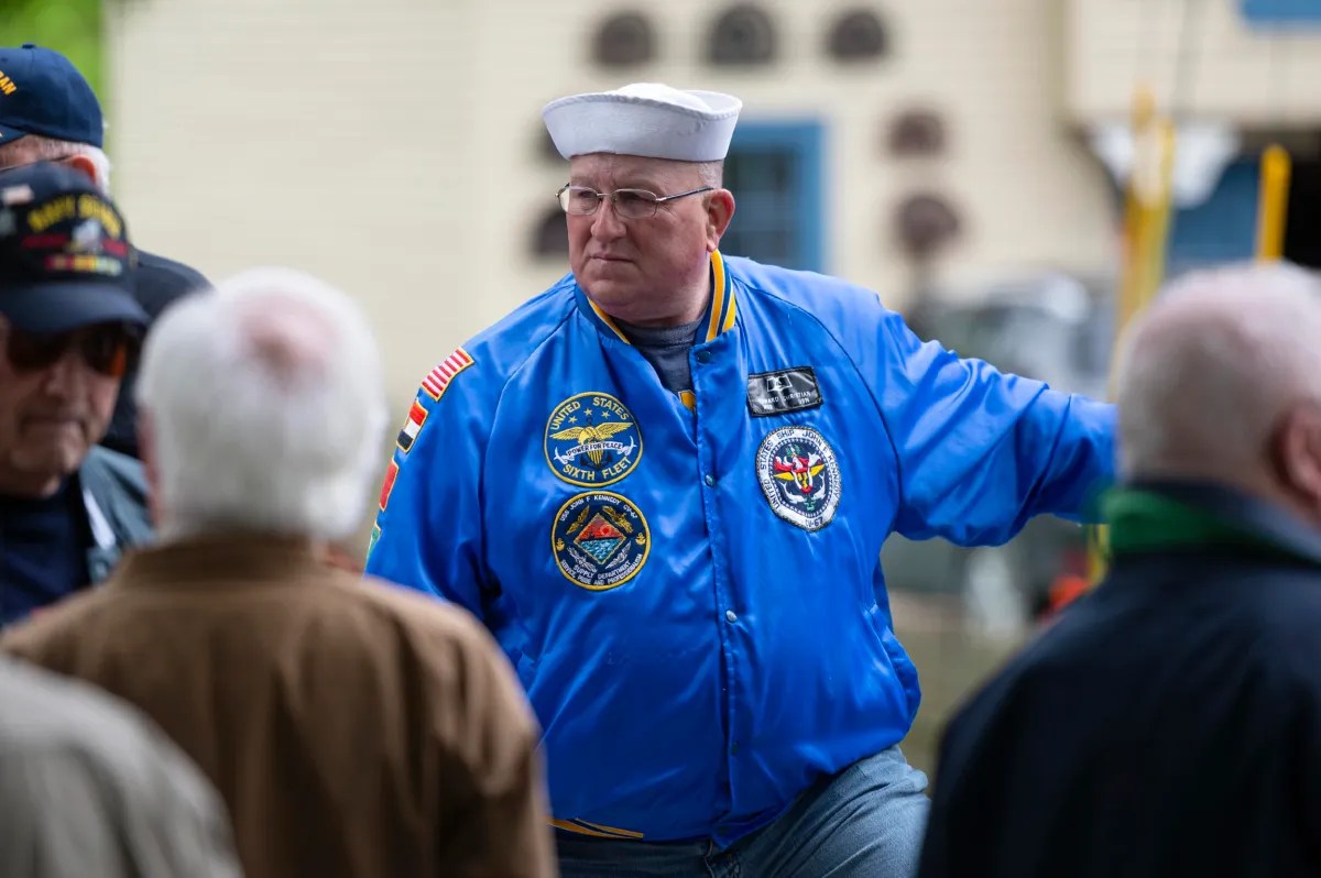 A man in a blue jacket with military patches and a white sailor hat stands among a group of people outdoors.