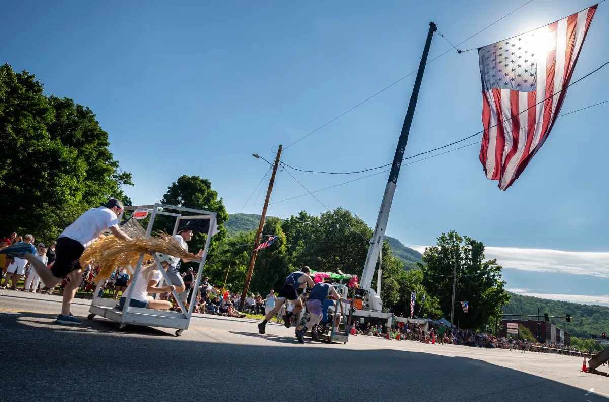 Participants in a homemade kart race push their vehicles down a street decorated with an American flag, while spectators watch under a clear blue sky.
