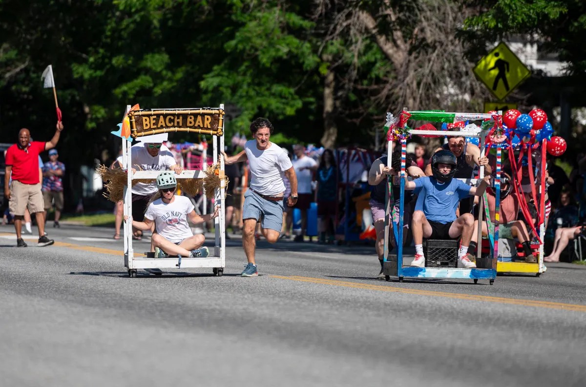 Participants in a soapbox race compete in creatively designed carts on a sunny day, with spectators watching and greenery in the background.