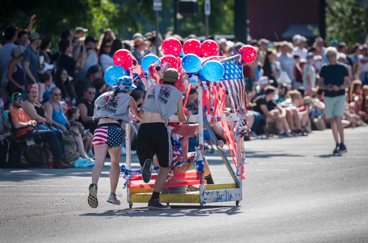 Participants in patriotic attire push a festive cart decorated with red, white, and blue balloons in a parade while spectators watch from the sidelines.