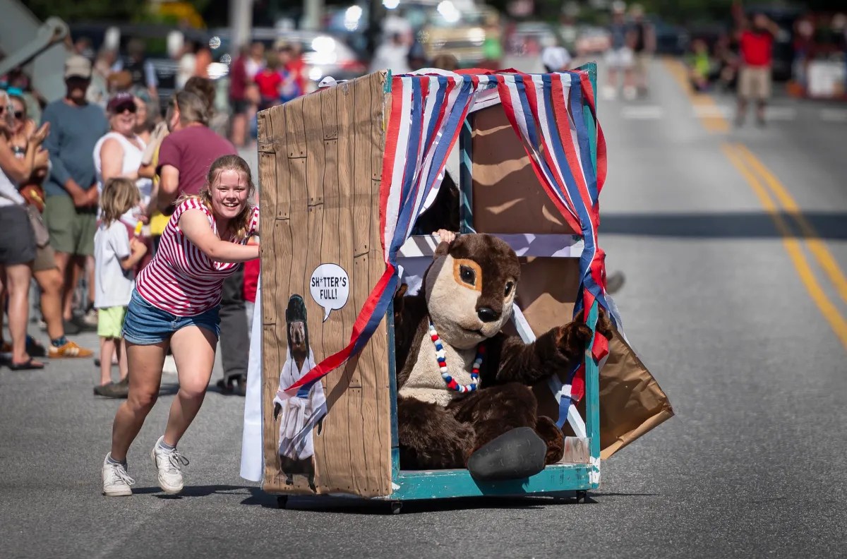 A person pushes a large, decorated booth with a person in a squirrel costume inside, along a street during a parade. Many spectators are observing on both sides of the road.