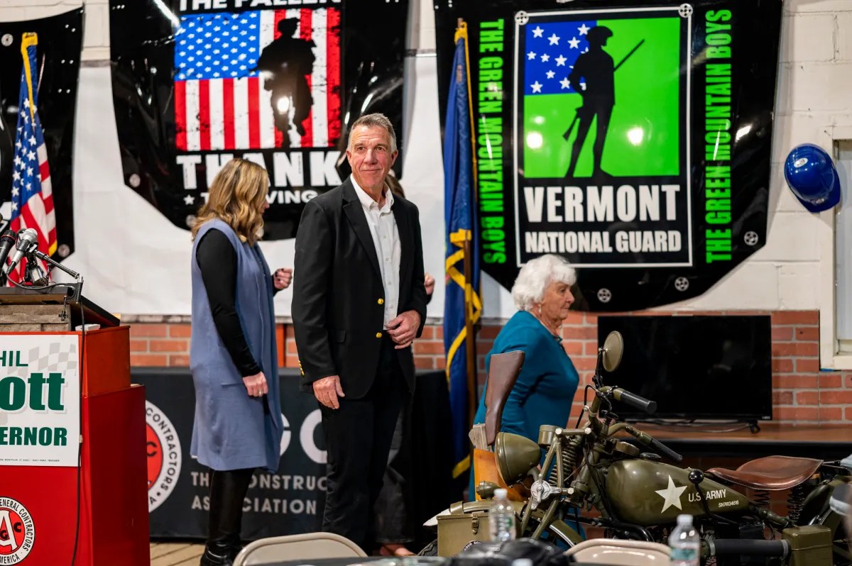 A man and two women stand near a podium in a room with military-themed banners and a vintage motorcycle.