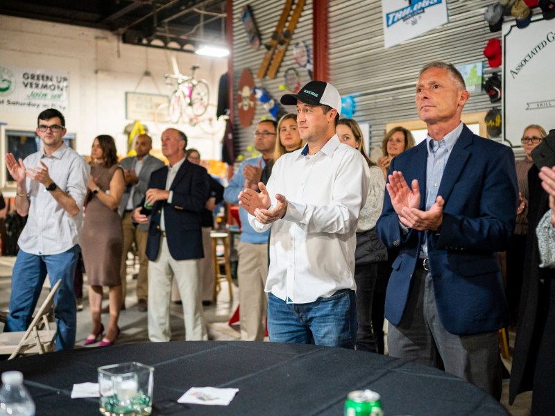 A group of people stands and applauds in a casual indoor setting with posters and bicycles on the walls.
