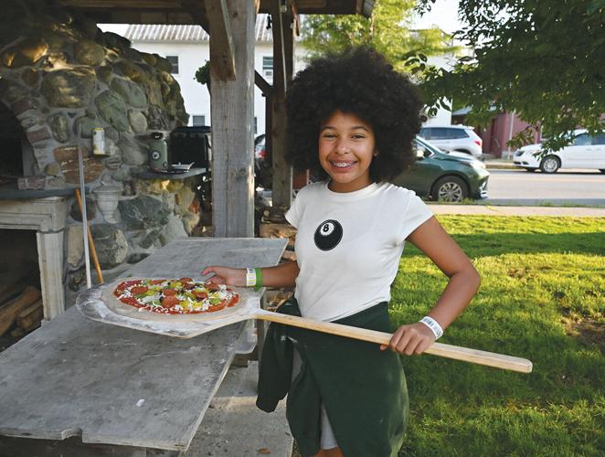 A young person stands outdoors holding a pizza peel with an uncooked pizza, smiling at the camera near a stone oven.
