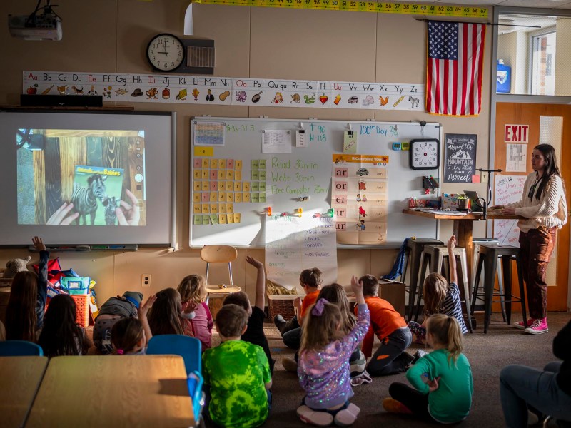 A teacher stands at the front of a classroom pointing at a projection screen, while children sit on the floor and at desks, some raising their hands. The American flag and educational materials are visible.