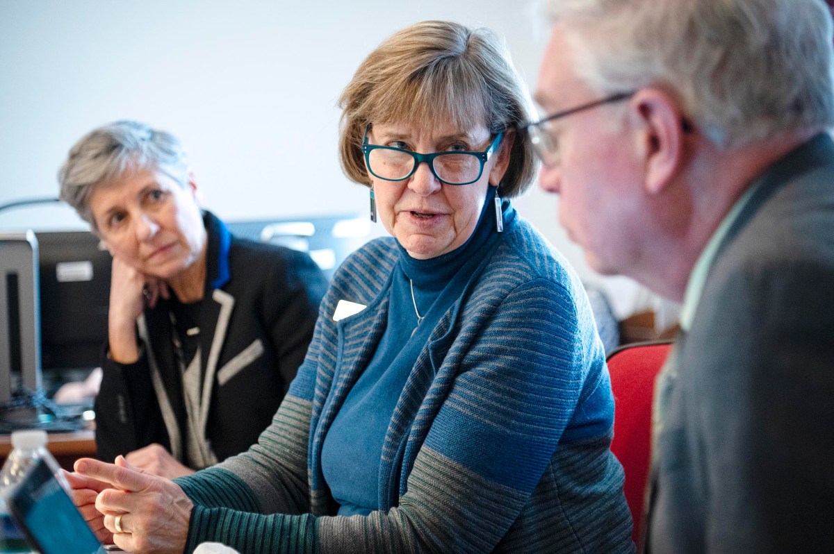 Three people engaged in conversation at a table in an office setting. The woman in the middle is wearing glasses and a blue striped sweater.