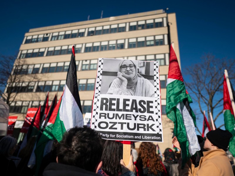 People holding Palestinian flags and a protest sign with a woman's photo read, "RELEASE RUMEYSA OZTURK," in front of a beige building.