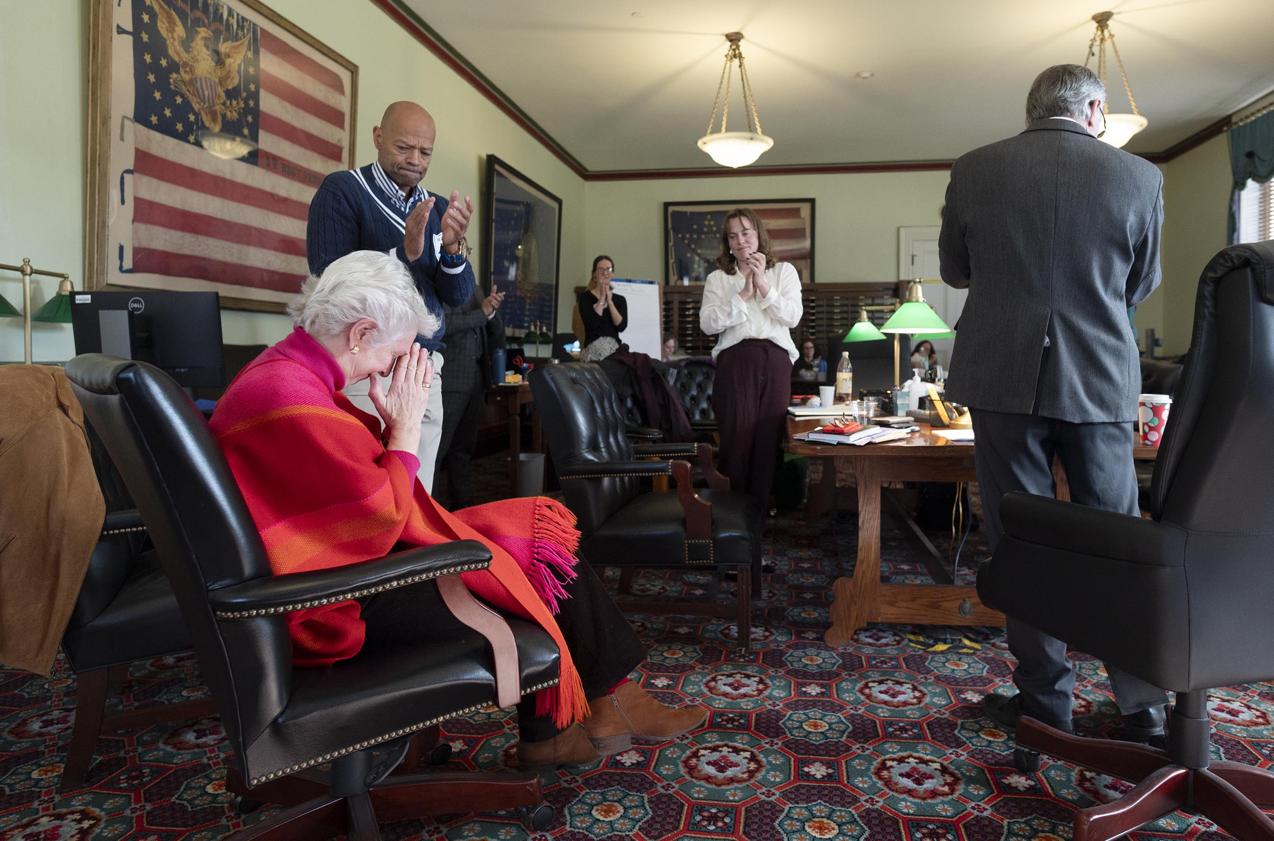A seated elderly woman is surrounded by standing people clapping in an office with flags on the wall.