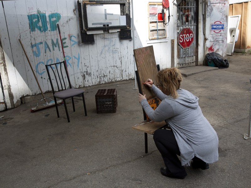 A person crouches while writing on a wooden board in an alley; two chairs, a crate, and graffiti reading "RIP James Carey" are visible nearby.