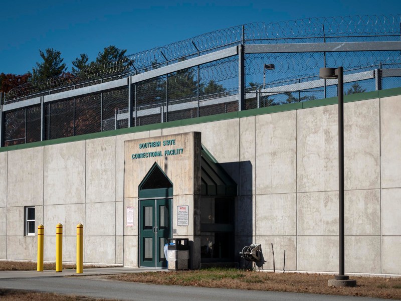 Exterior of a correctional facility showing a concrete wall topped with barbed wire, a sign at the entrance, and security cameras.
