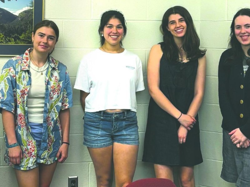Six people stand indoors against a white wall, some smiling, dressed in casual and semi-formal clothing. A framed photo and outlet are visible in the background.