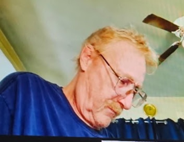 A man with glasses and a mustache, wearing a blue shirt, looks down in a room with a ceiling fan and dark curtains in the background.