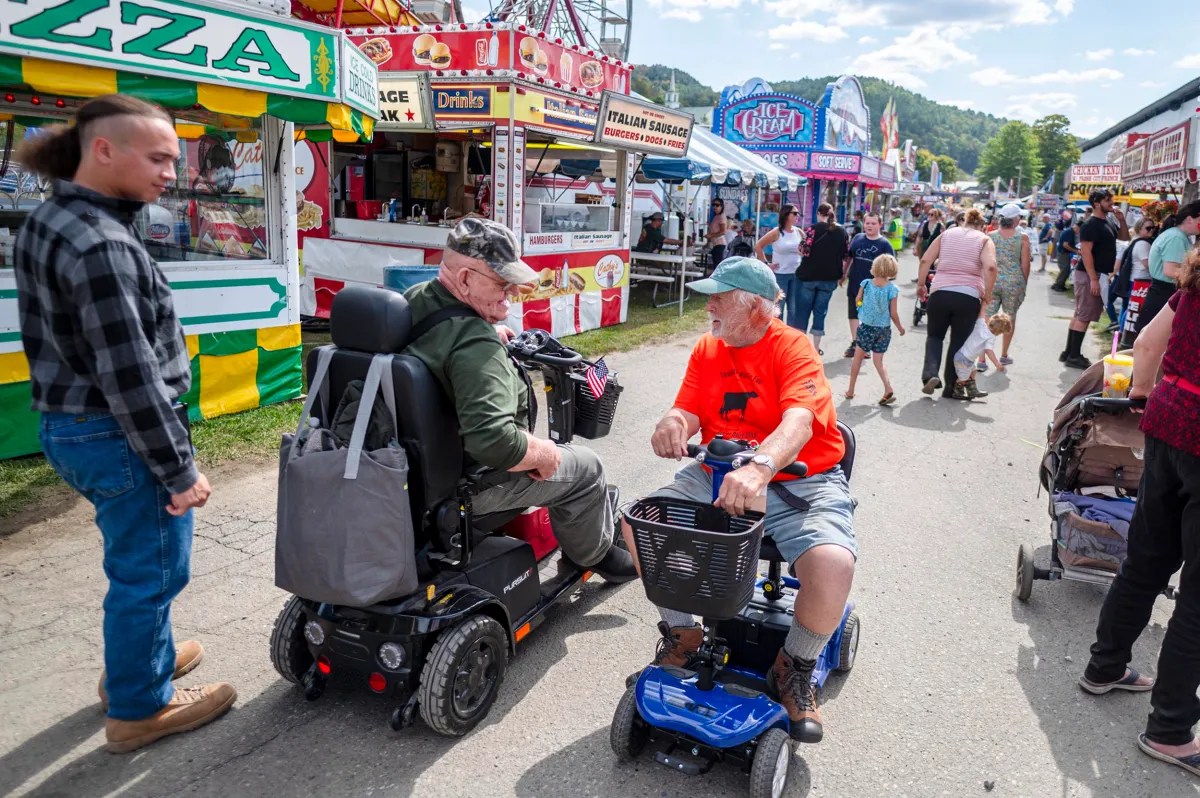 Two men in mobility scooters talk at an outdoor fair with food stalls, families, and crowds visible in the background on a sunny day.
