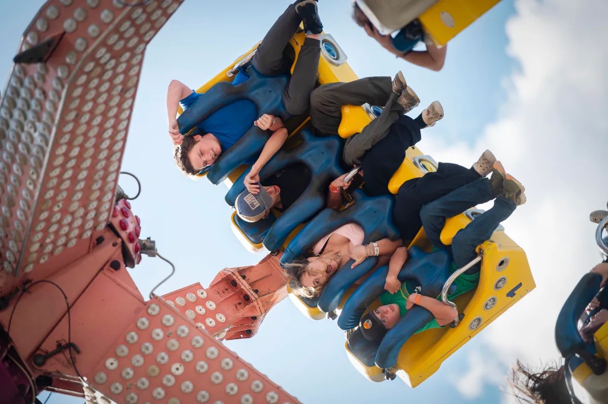 A group of people ride an amusement park attraction, suspended upside down in blue and yellow seats against a bright sky.