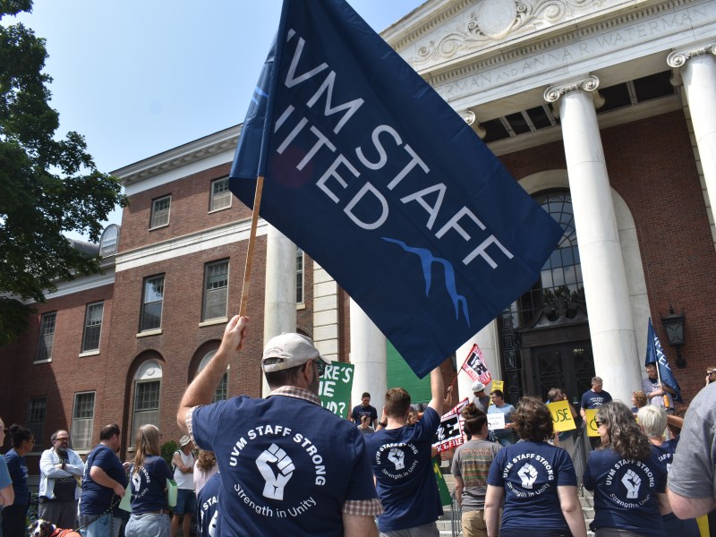 People gather outside a brick building with columns, holding signs and a large "UVM Staff United" flag at a rally. Many wear matching "UVM Staff Strong" shirts.