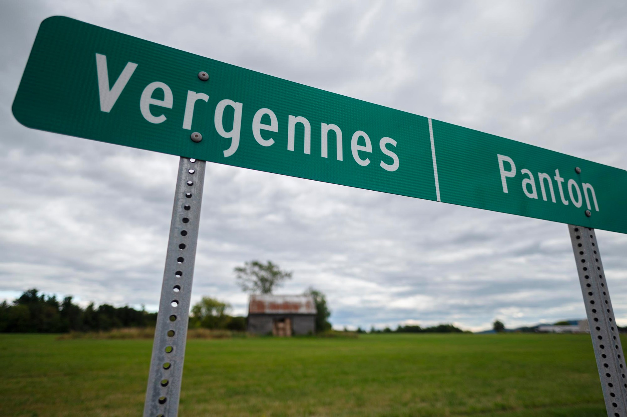 A green road sign with white text showing directions to Vergennes and Panton stands in front of a grassy field and an overcast sky.