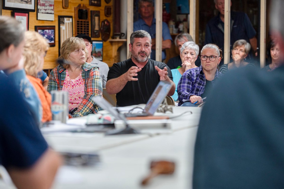 A man speaks to a group of people seated around a table during a meeting in a room with wooden walls and framed items.
