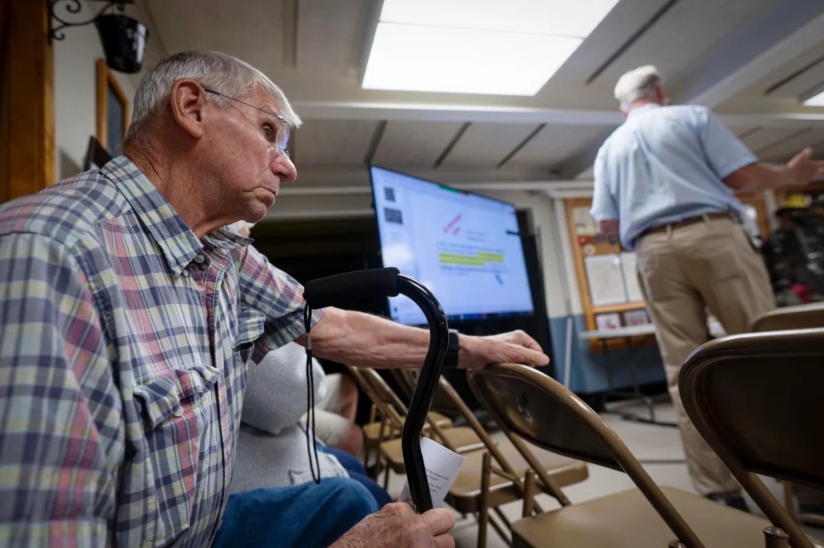 An older man with a cane sits in a room with folding chairs, looking at a presentation screen, while another man stands in the background.