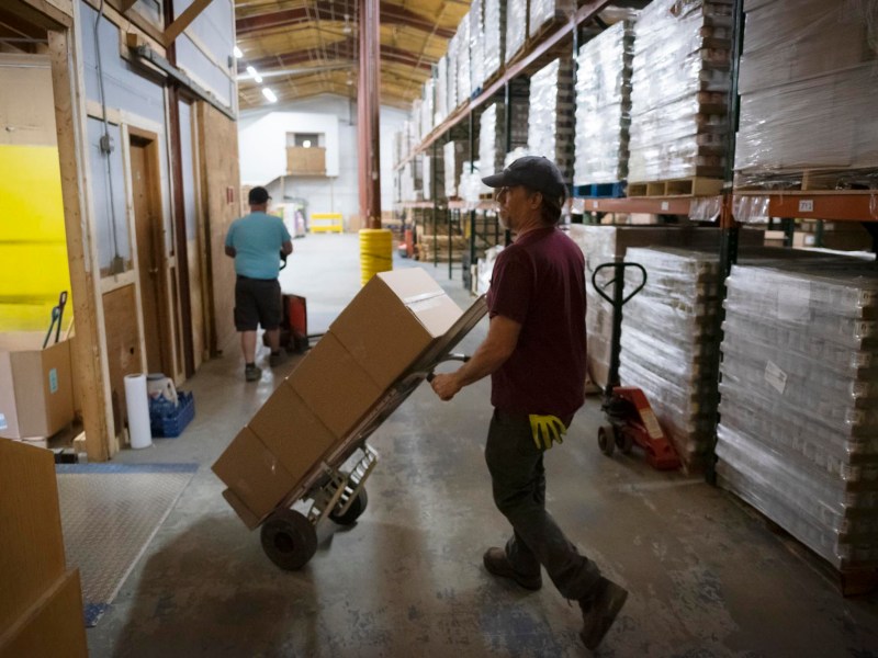 Two workers move boxes on dollies inside a warehouse lined with shelves holding stacked pallets of goods.