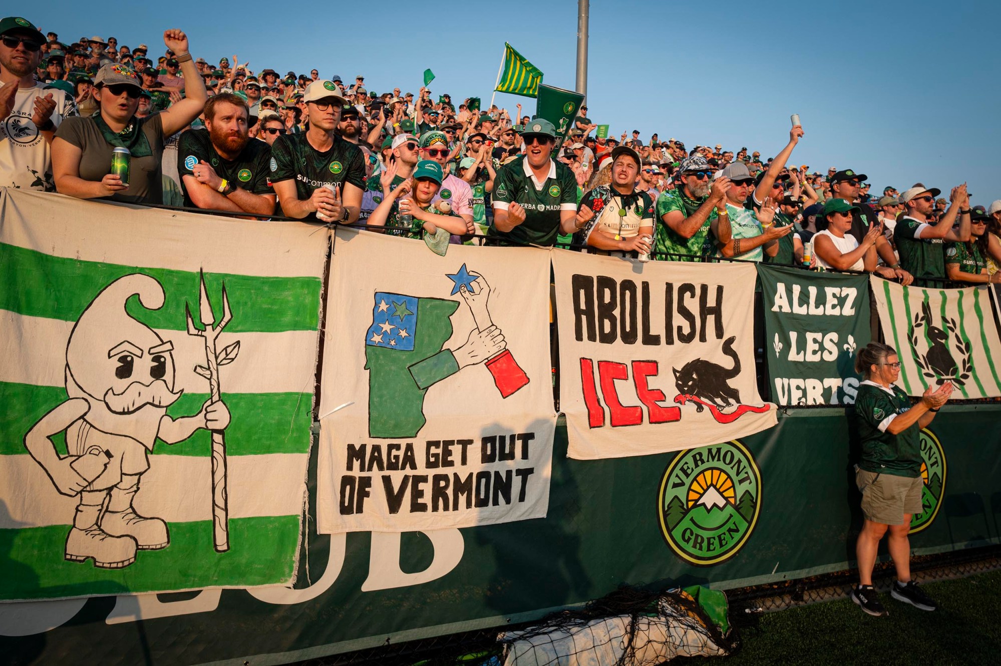 A crowd of soccer fans stands behind banners, including images and slogans such as “MAGA Get Out of Vermont,” “Abolish ICE,” and a Vermont Green FC logo.