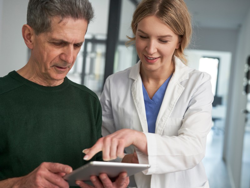 A healthcare professional in a white coat shows information on a tablet to an older man in a green sweater.