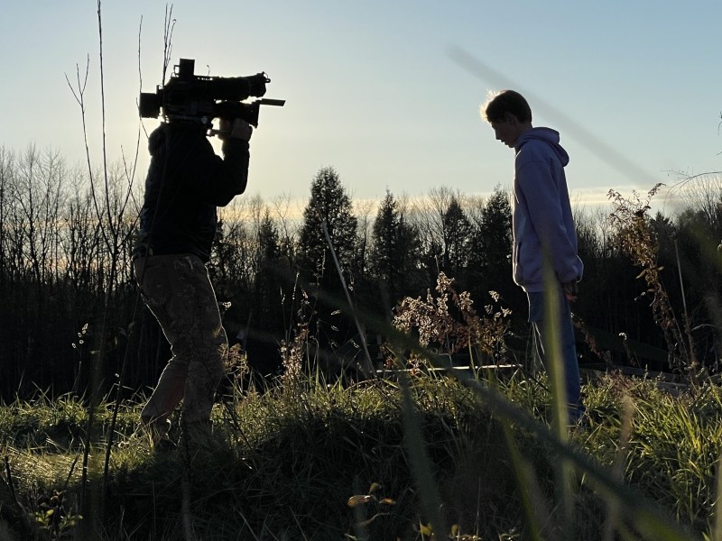 A person operates a large video camera, filming another person who is standing on grass in an outdoor setting during sunset.
