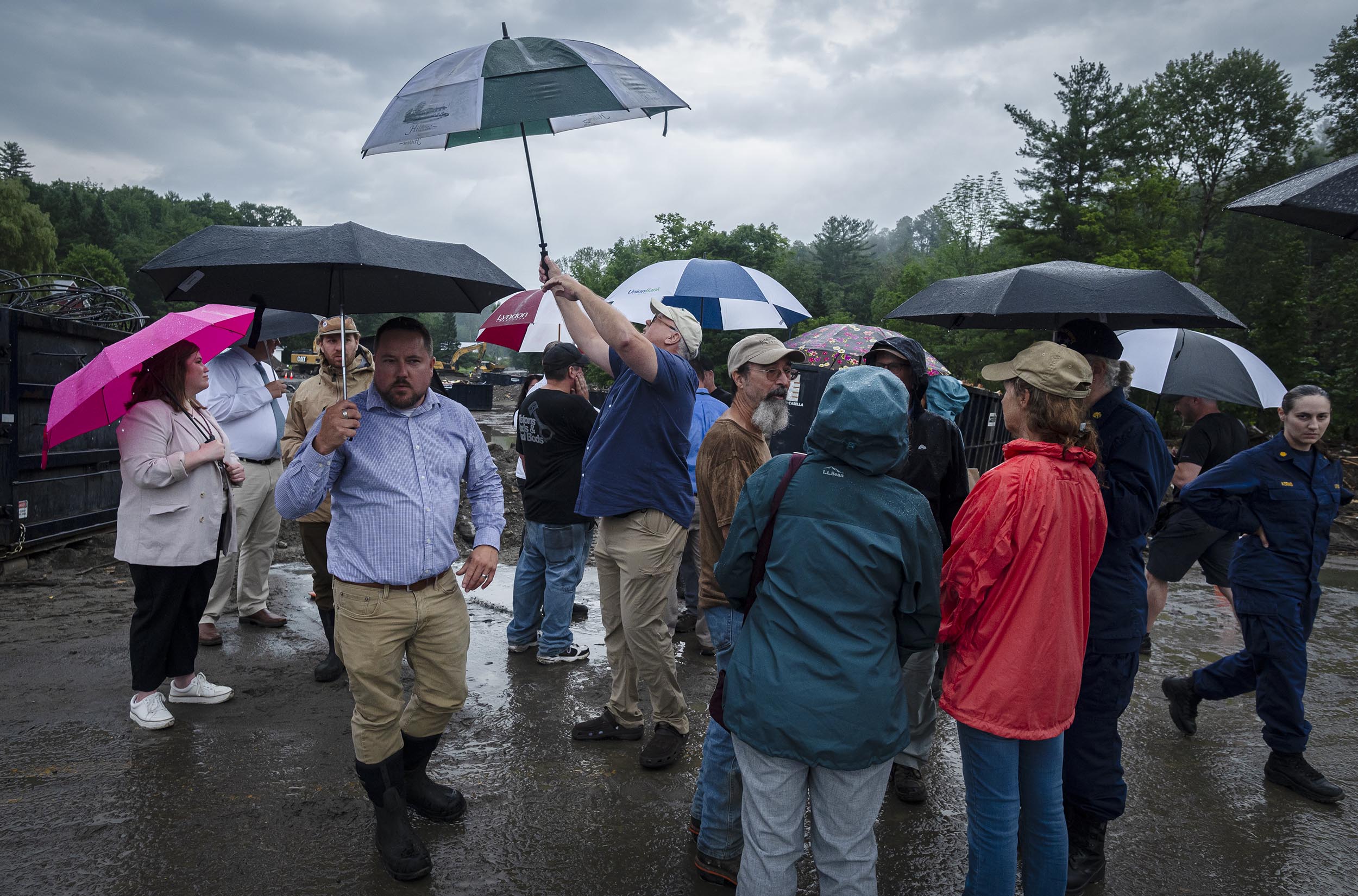 People stand outside in the rain, holding umbrellas and wearing jackets. The ground is wet and muddy. Trees and overcast sky are visible in the background.