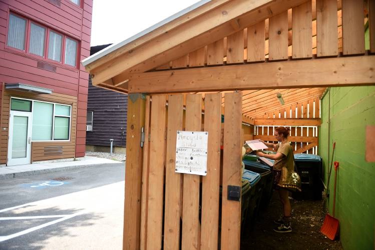 A person places items into bins inside a wooden recycling enclosure next to a building, with a posted recycling schedule visible on the fence.
