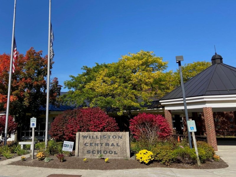 A building with a sign reading "Williston Central School" is surrounded by trees and flowers. Two flagpoles, one with an American flag, are on the left side.