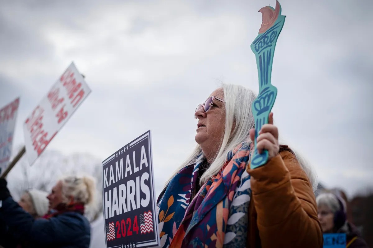 A woman holds a torch cutout and a "Kamala Harris 2024" sign at a gathering. Others in the background hold protest signs.