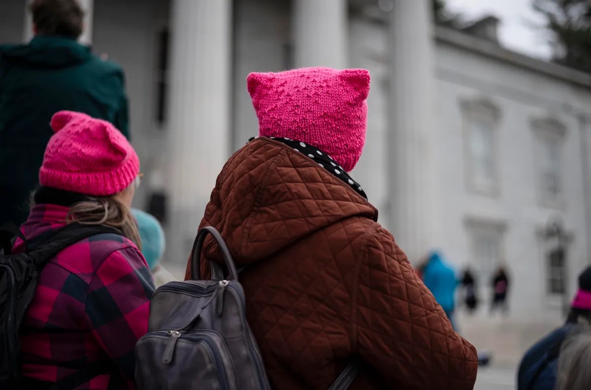 Individuals wearing pink knitted hats and winter coats stand outside a building with tall white columns.