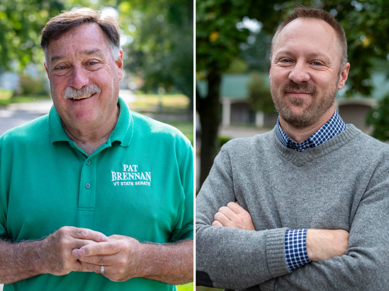 Two men are posing outdoors. One wears a green polo with text and the other wears a gray sweater over a checkered shirt. Both smile and stand in a tree-lined setting.