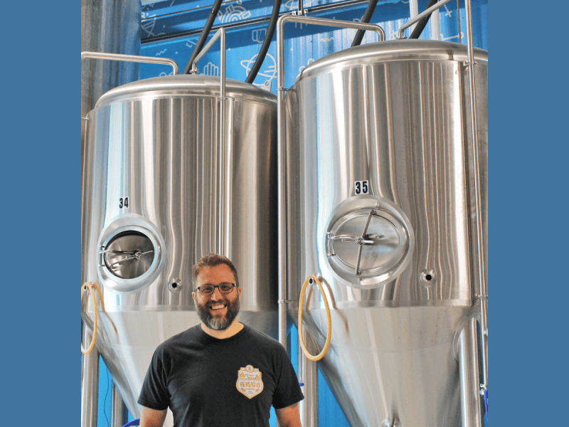 A man with a beard and glasses stands smiling in front of two large stainless steel brewing tanks labeled 34 and 35.