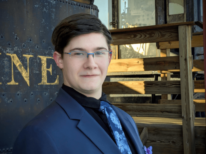 A young man in a dark suit, glasses, and a blue patterned tie stands in front of a wooden staircase and a black metal structure with gold lettering.